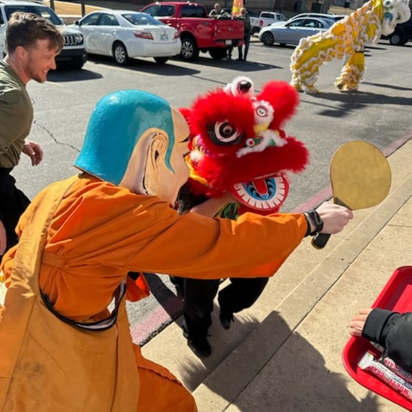 A person in an orange robe and blue mask leads a red lion dance costume with a paddle.