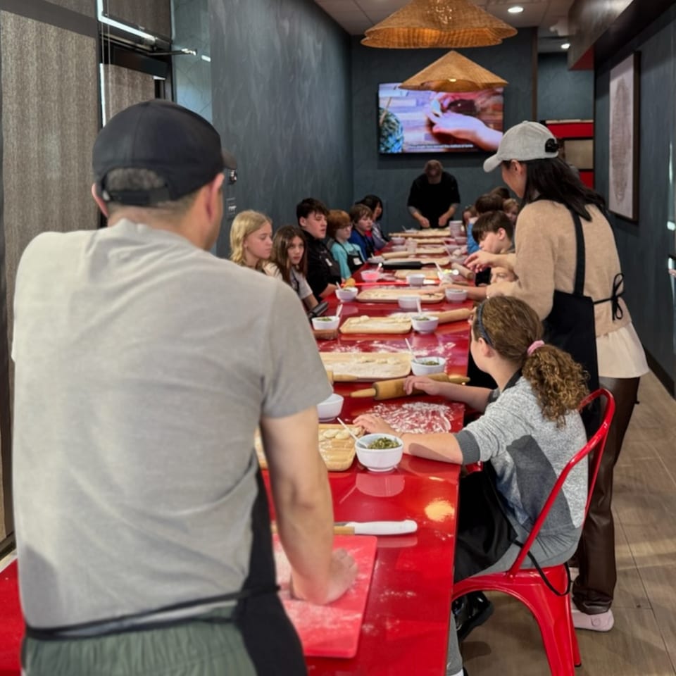 Children and adults learning to make dumplings at a long red table in Red Lotus Noodle House.